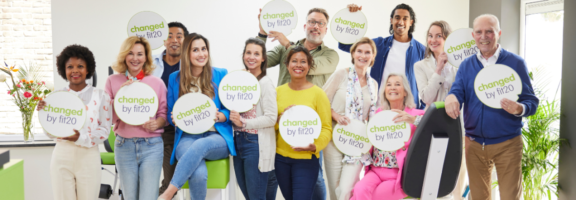 Groep mensen met changed by fit20-borden, in een lichte ruimte met sportapparatuur.